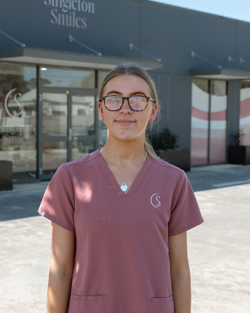 Amber, dental assistant at Singleton Smiles, smiling warmly in pink scrubs seated in a modern dental practice