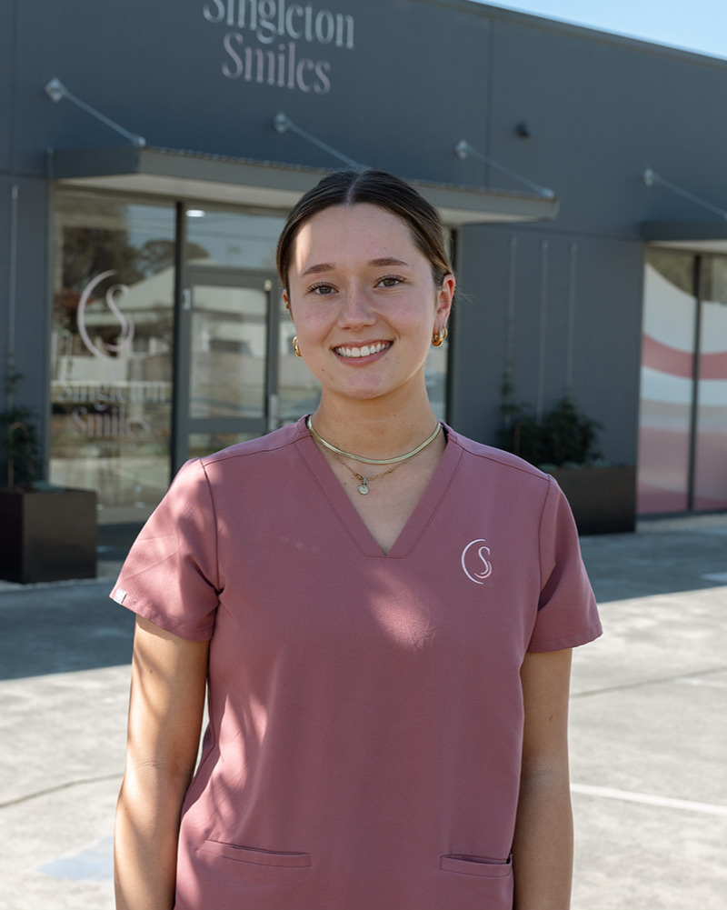 Amelia (Milly), dental assistant at Singleton Smiles, smiling warmly in pink scrubs seated in a modern dental practice