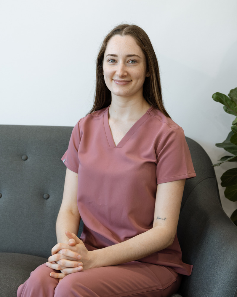 Brodie, dental assistant at Singleton Smiles, smiling warmly in pink scrubs seated on a grey couch