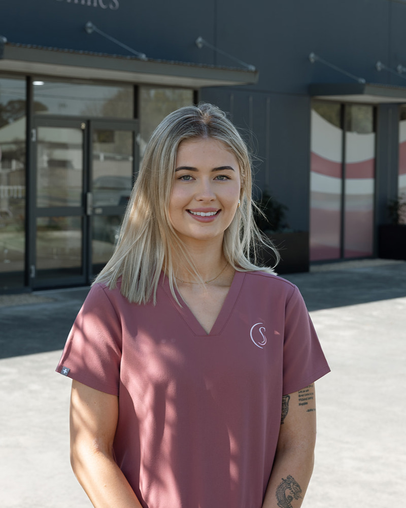 Lily, dental assistant at Singleton Smiles, smiling warmly in pink scrubs seated in a modern dental practice setting.