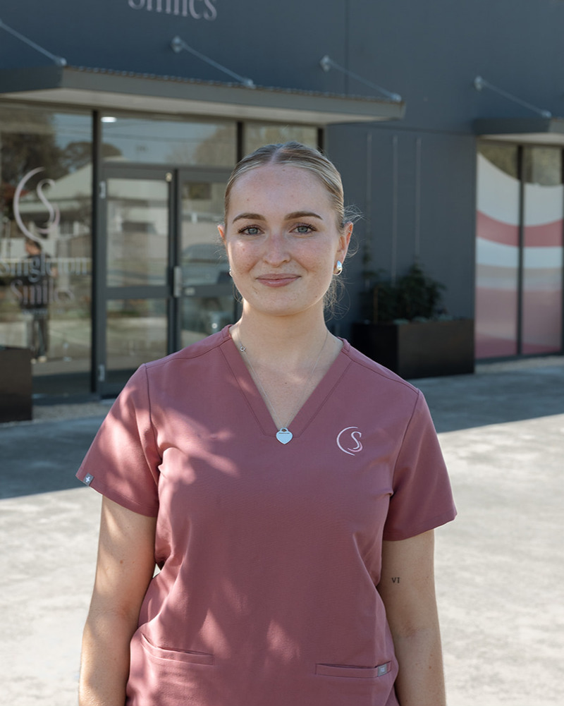 Madison, dental assistant at Singleton Smiles, smiling warmly in pink scrubs seated in a modern dental practice